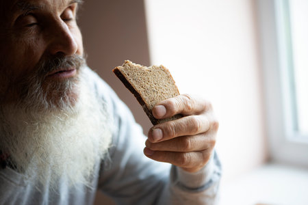 Old sad man with a long gray beard sitting by the table and eating soup and bread