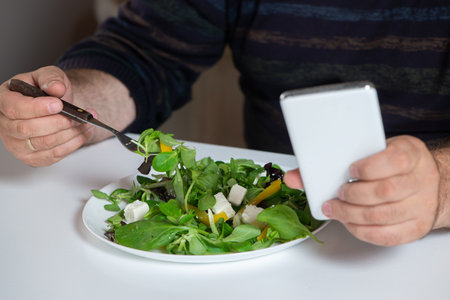 Mature  man sits at table, eating salad greens and using a smartphone.