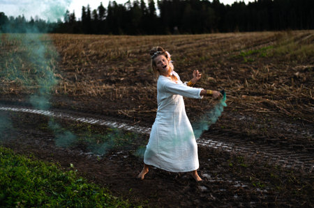 Happy woman in white dress with green smoke bomb dancing on muddy fieldの写真素材