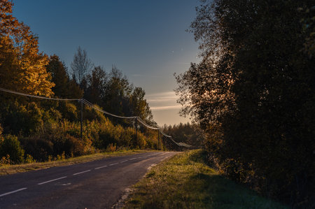 Soft light of the setting evening sun reflects on power lines along an asphalt country road, golden hour vibes in Latviaの写真素材
