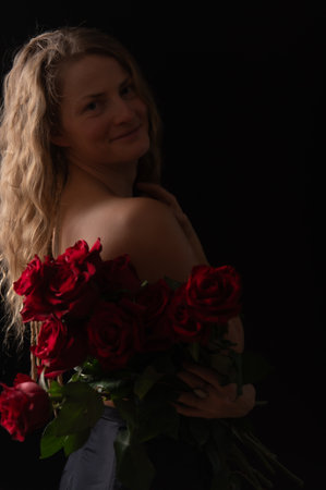Beautiful woman with bare shoulders and red roses on a dark studio background. Beauty studio portrait. Sensual model with red roses flowers, isolated on black. Charming mature natural female without makeup.の写真素材