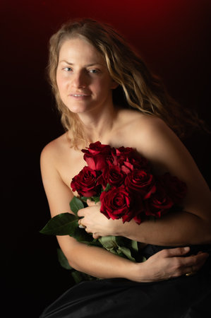 Beautiful woman with bare shoulders and red roses on a dark studio background. Beauty studio portrait. Sensual model with red roses flowers, isolated on black. Charming mature natural female without makeup.の写真素材