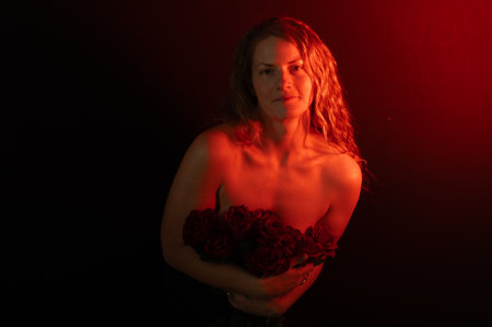 Beautiful woman with bare shoulders and red roses on a dark studio background. Beauty studio portrait. Sensual model with red roses flowers, isolated on black. Charming mature natural female without makeup.の写真素材