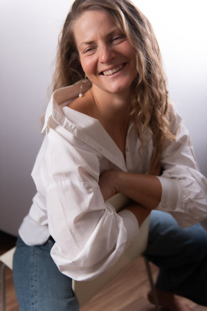 Beautiful woman in white shirt posing in studio on chair. Happy adult female with long curly hair on white studio background. Expressive portrait with natural emotions.の写真素材