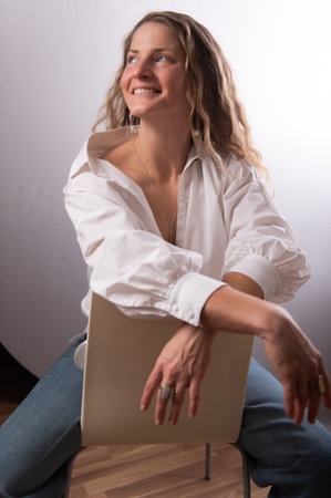 Beautiful woman in white shirt posing in studio on chair. Happy adult female with long curly hair on white studio background. Expressive portrait with natural emotions.の写真素材