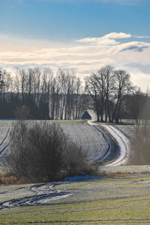 A snowy winding path leads through frosty fields toward a distant barn in Latvia, framed by bare trees and a bright winter sky.の写真素材