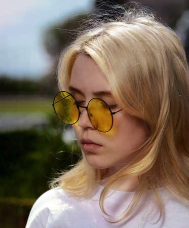 A young blonde girl outdoors in summer in yellow round glasses closed her eyes thoughtfully. Soft focusの写真素材