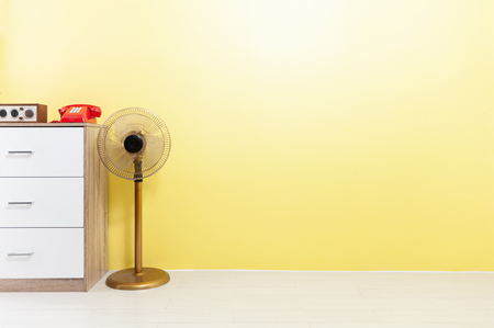 Old fan beside wooden drawer, red telephone and classic radio on yellow background and wooden floor.の写真素材