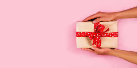 Woman hands holding wrapped craft paper box with gift and decorated with red bow on pink background....