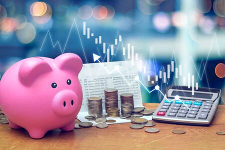 Coins stack on saving account book from bank with pink piggy bank and calculator on wooden table with candlestick chart suitable for finance and investment concept and blue bokeh background.の写真素材