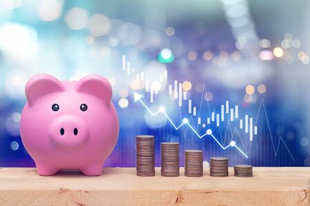 Pink piggy bank and coins stack on wooden table with candlestick chart suitable for finance and investment concept and blue bokeh background.の写真素材