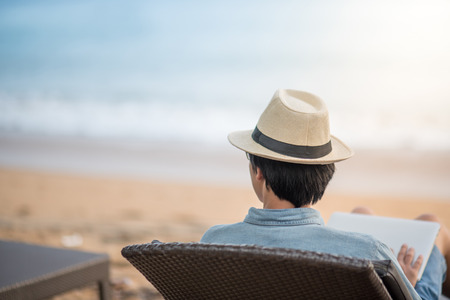 Young asian man sitting on bench at tropical beach and looking to the sea for relaxation after work, digital nomad lifestyle or working outdoor conceptsの写真素材