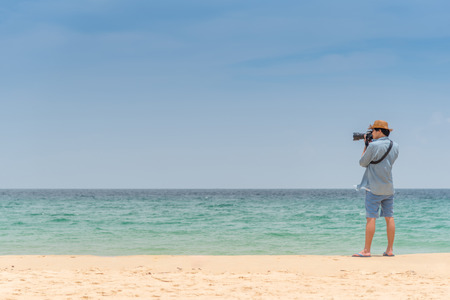 young Asian man photographer with jean shirt and hat take photo of tropical island beach and turquoise sea, seascape background for summer holiday and vacation travel conceptsの写真素材