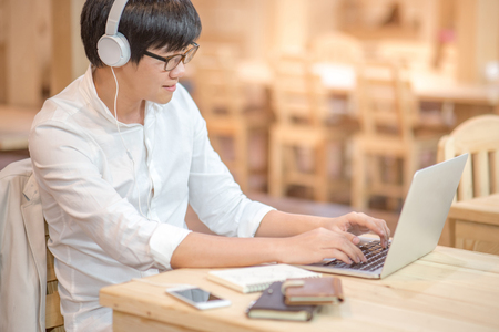 Asian happy young man with headphones listening to music and working on modern laptop computer in vintage interior workplace, gadget lifestyle and online learning conceptsの写真素材