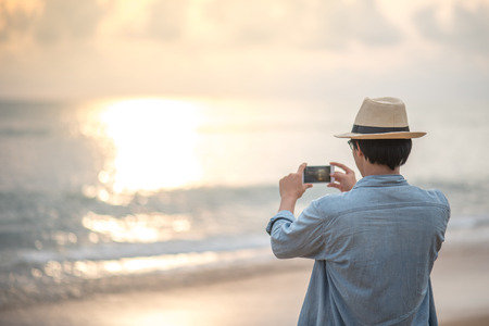 young Asian happy man with jean shirt and hat taking photos sunset on tropical beach island by smartphone, summer holiday or vacation travel conceptsの写真素材