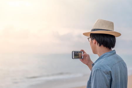 young Asian happy man with jean shirt and hat taking photos sunset on tropical beach island by smartphone, summer holiday or vacation travel conceptsの写真素材
