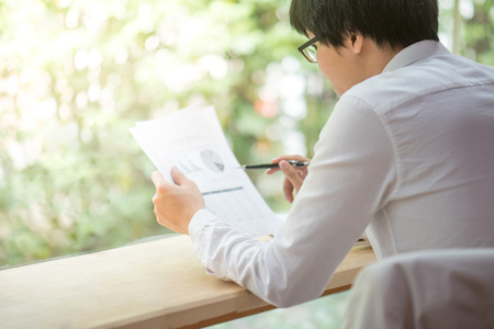 young Asian business man checking summary report document for analysis, smart working and good potential management in office workspaceの写真素材