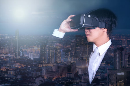 young asian business man wearing suit playing virtual reality goggles with night city view background. smartphone using with VR headset. technology and innovation conceptsの写真素材