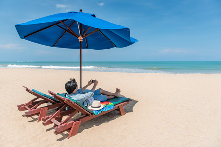 young man lying on wooden beach bench with blue umbrella and take photo of turquoise sea and beautiful sky by smartphone, vacation time and summer holiday concepts, digital nomad lifestyleの写真素材