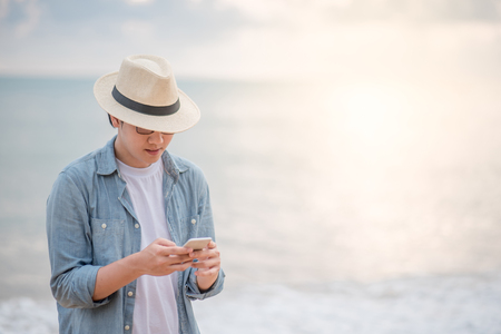 Young Asian man with jean shirt and hat holding smartphone and checking social media application on tropical beach at sunset, background for summer holiday vacation or global communication conceptsの写真素材