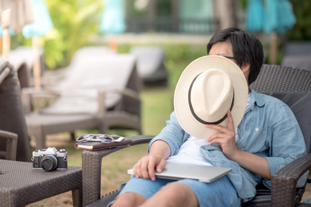 Young Asian man lying on bench near the beach with laptop, notebook and camera for outdoor working, digital nomad lifestyle conceptsの写真素材