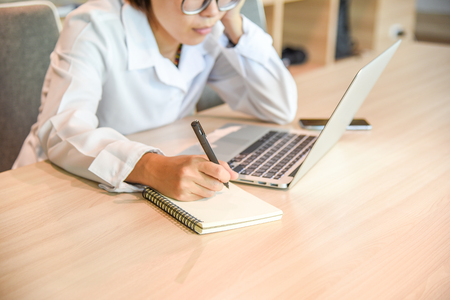 Businesswoman in workplace writing on blank notebook and using laptop on wooden office desk for business information management and contact customer with online social media technologyの写真素材