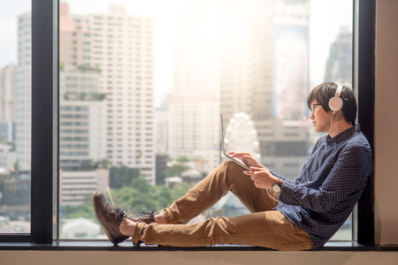 Young asian man relaxing enjoy watching movie from his laptop computer with headphones on glass window. Urban lifestyle in living space.の写真素材