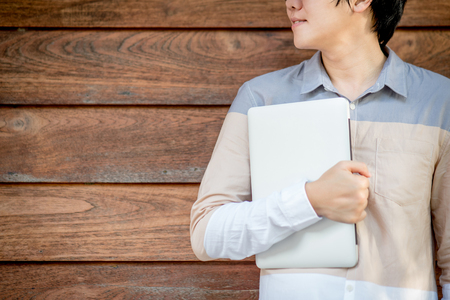 Young Asian university student man holding laptop computer with brown wood wall in background, education and college lifestyle conceptsの写真素材
