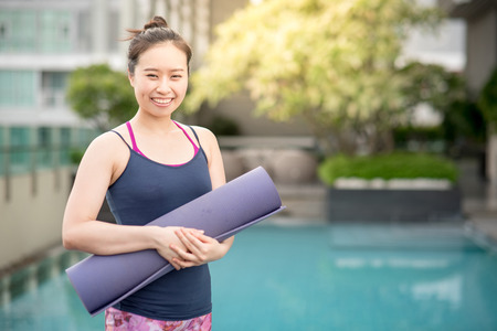 Young Asian happy woman holding purple yoga mat at swimming pool, outdoor exercise and healthy lifestyle conceptsの写真素材