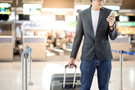 Young asian man with his suitcase luggage using smartphone while waiting for airline flight in the international airport terminal, business travel and online check in conceptsの写真素材