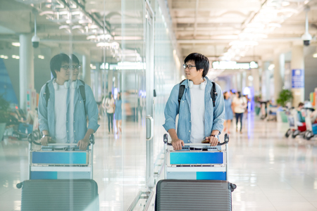 Young Asian man walking with airport trolley and his suitcase luggage in the international airport terminal, arrival from travel abroadの写真素材