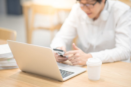 Young Asian businessman using smart phone and working with laptop computer in coffee shop. Freelance job and IT modern lifestyle, work life balance conceptの写真素材