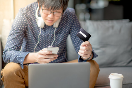 Young Asian man half face holding credit card and using smartphone for online shopping in his workspace, urban lifestyle conceptの写真素材