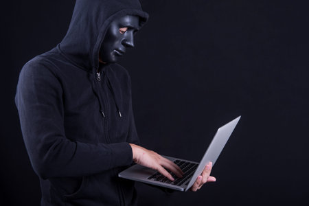 Anonymous male hacker using black mask to cover his face carrying laptop computer. Internet security...