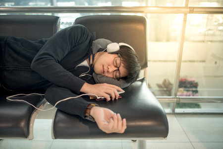 Young Asian man with eyeglasses and headphones sleeping on bench while waiting for connecting flight...