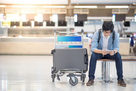 young asian guy using smartphone while waiting for check in and drop his suitcase luggage at airline check-in counter of international airport terminalの写真素材