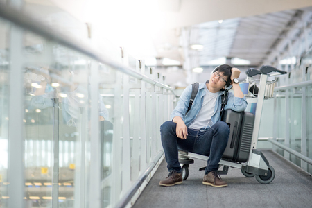 Young Asian man sitting on airport trolley with his suitcase luggage in the international airport terminal, waiting for travel abroadの写真素材