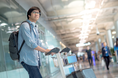 Young Asian man walking with airport trolley and his suitcase luggage in the international airport terminal, arrival from travel abroadの写真素材