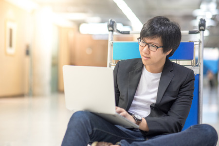 Young asian man working on airport trolley with his laptop computer during waiting for a connecting flight, freelance lifestyle and digital nomad conceptsの写真素材
