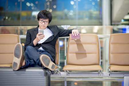 Young Asian well-dressed businessman using smartphone sitting on bench near his suitcase luggage while waiting for connecting flight in airport terminal. man in business travel conceptの写真素材