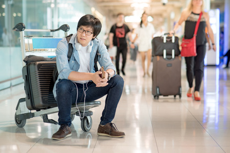 Young Asian man sitting on airport trolley with his suitcase luggage in the international airport terminal, waiting for travel abroadの写真素材