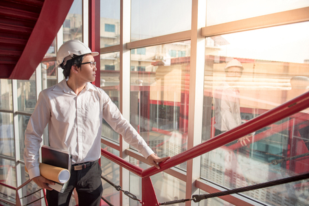 Young Asian Engineer or Architect holding files and architectural drawing wearing personal protective equipment safety helmet at construction site. Civil engineering, Architecture and building conceptの写真素材