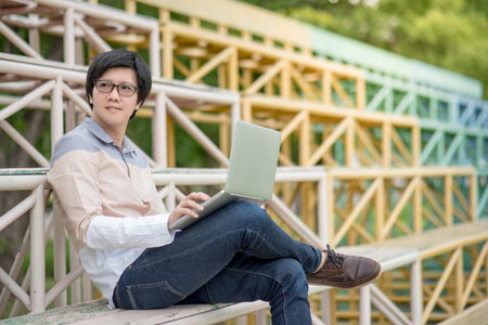 Young Asian student man sitting on grandstand and using laptop computer, university lifestyle in...