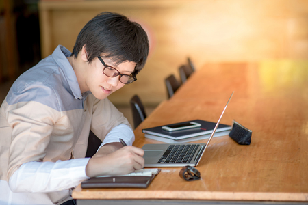 Young Asian man writing on notebook and working with laptop computer in college building. high school or university student, educational conceptsの写真素材