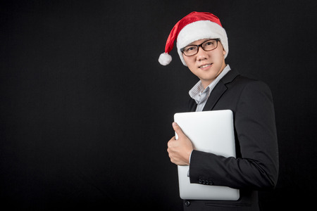 Young Asian businessman wearing suit and santa hat holding laptop computer on black background, male entrepreneur in christmas holiday and new year party conceptsの写真素材