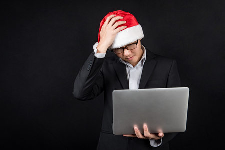 Young Asian businessman wearing suit and santa hat using laptop computer on black background, male entrepreneur in christmas holiday and new year party conceptsの写真素材