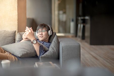 Young Asian man using smartphone for listening to music, taking a break and relaxing on sofa in living space, high school or university college student, educational conceptの写真素材