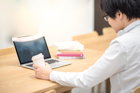 Young Asian man holding coffee cup while working with laptop computer. Male university student doing education research in library.の写真素材