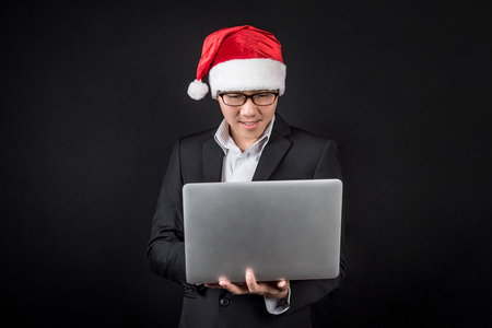 Young Asian businessman wearing suit and santa hat using laptop computer on black background, male entrepreneur in christmas holiday and new year party conceptsの写真素材