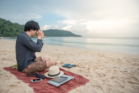 Young Asian man taking photo while sitting on tropical beach, digital nomad or freelance lifestyle conceptsの写真素材